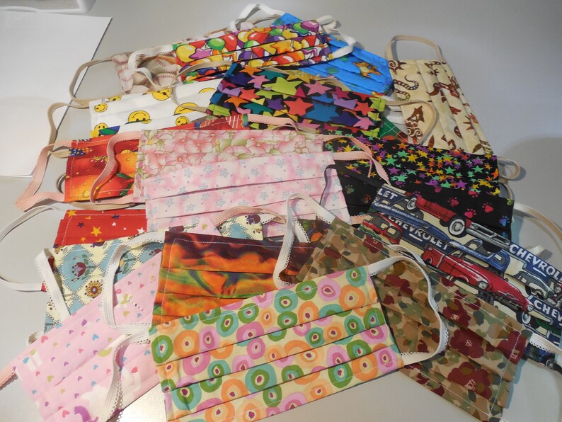 May include: A collection of assorted fabric face masks with elastic ear loops. The masks feature various patterns, including stars, smiley faces, floral prints, and car designs. Colors include red, blue, pink, and green.