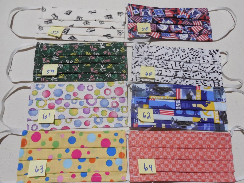 May include: Eight different fabric face masks with various patterns, including musical notes, circles, and American flags. Each mask has a yellow sticky note with a number on it, ranging from 57 to 64.