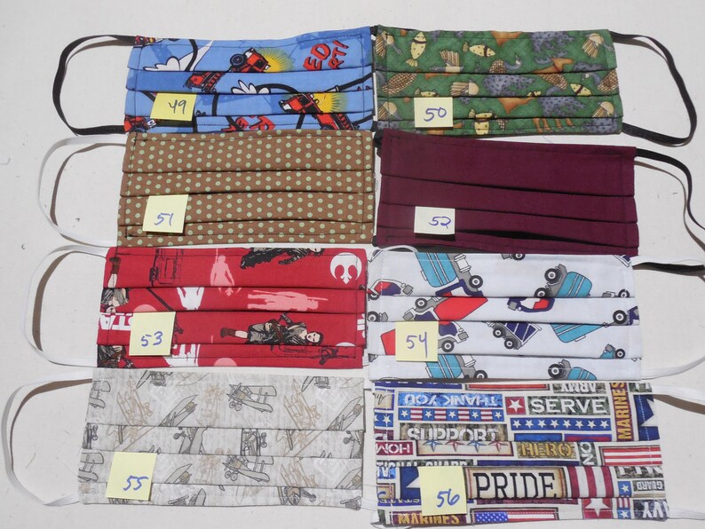 May include: Six different fabric face masks with various patterns. The masks are pleated and have elastic ear loops. The patterns include a blue and red fire truck design, a green and brown animal print, a brown with white polka dots, a maroon solid color, a red with white Star Wars characters, a white with blue trucks, a gray with brown airplanes, and a red, white, and blue patriotic design. The masks are numbered 49 through 56.