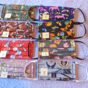 May include: Eight assorted fabric face masks with different patterns, including footballs, unicorns, owls, and military aircraft. Each mask has elastic ear loops and a pleated design. Numbers 1-8 are on small tags attached to each mask.