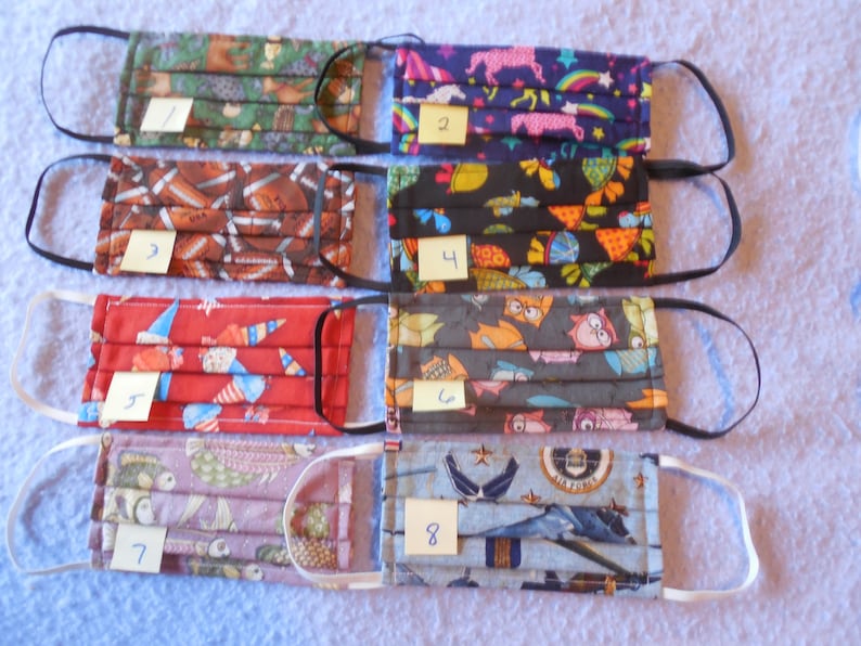 May include: Eight assorted fabric face masks with different patterns, including footballs, unicorns, owls, and military aircraft. Each mask has elastic ear loops and a pleated design. Numbers 1-8 are on small tags attached to each mask.