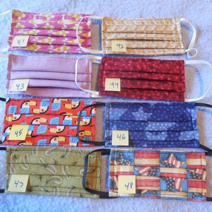 May include: Assortment of handmade face masks with various patterns, including fish, swirls, solid lavender, red stars, toucans, blue stars, feathers, and American flags. Each mask has elastic ear loops and a number tag.