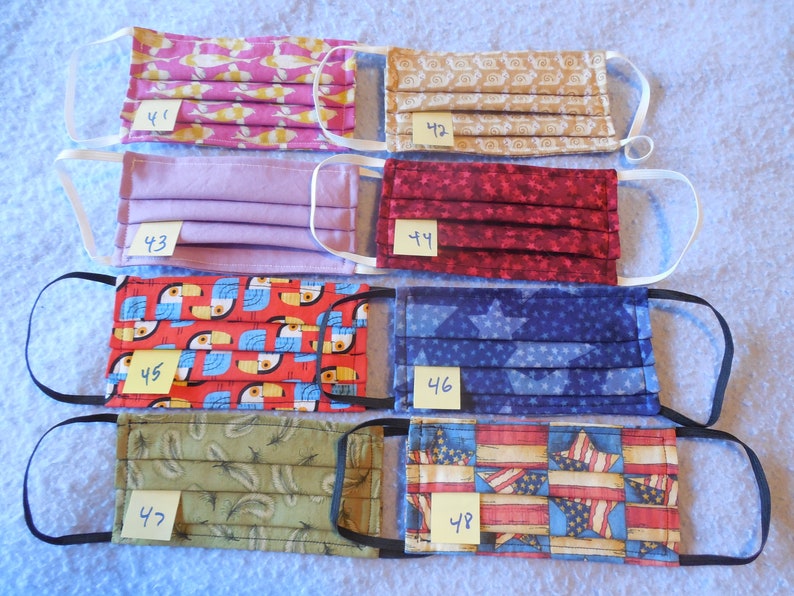 May include: Assortment of handmade face masks with various patterns, including fish, swirls, solid lavender, red stars, toucans, blue stars, feathers, and American flags. Each mask has elastic ear loops and a number tag.