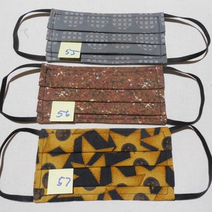 May include: Three pleated face masks with black elastic ear loops. The top mask is gray with a blue dot pattern, the middle mask is brown with a white floral pattern, and the bottom mask is yellow and black with a geometric design. Each mask has a small tag with a number.