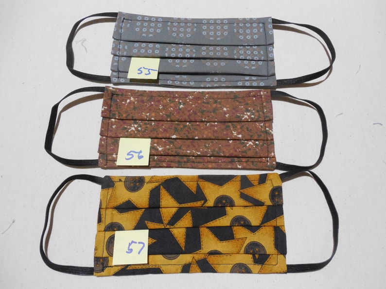 May include: Three pleated face masks with black elastic ear loops. The top mask is gray with a blue dot pattern, the middle mask is brown with a white floral pattern, and the bottom mask is yellow and black with a geometric design. Each mask has a small tag with a number.