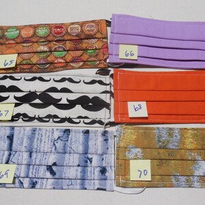 May include: Five different fabric face masks with numbers 65, 66, 67, 68, and 70 written on sticky notes. The masks are pleated and have different patterns, including bottle caps, mustaches, wood grain, and abstract designs.