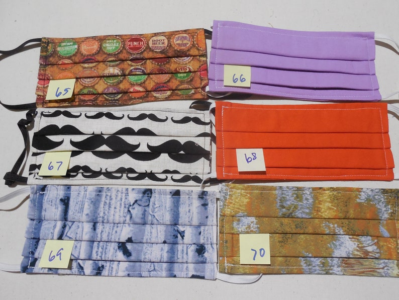 May include: Five different fabric face masks with numbers 65, 66, 67, 68, and 70 written on sticky notes. The masks are pleated and have different patterns, including bottle caps, mustaches, wood grain, and abstract designs.
