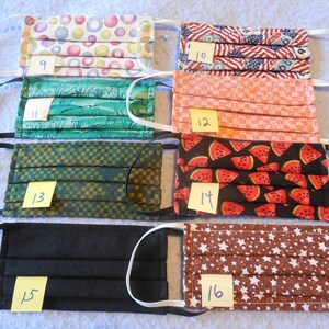 May include: Assortment of pleated face masks with elastic ear loops. Patterns include colorful circles, American flags, a green landscape, a peach-colored grid, watermelon slices, a black solid, and brown stars. Each mask has a number on a yellow sticky note.