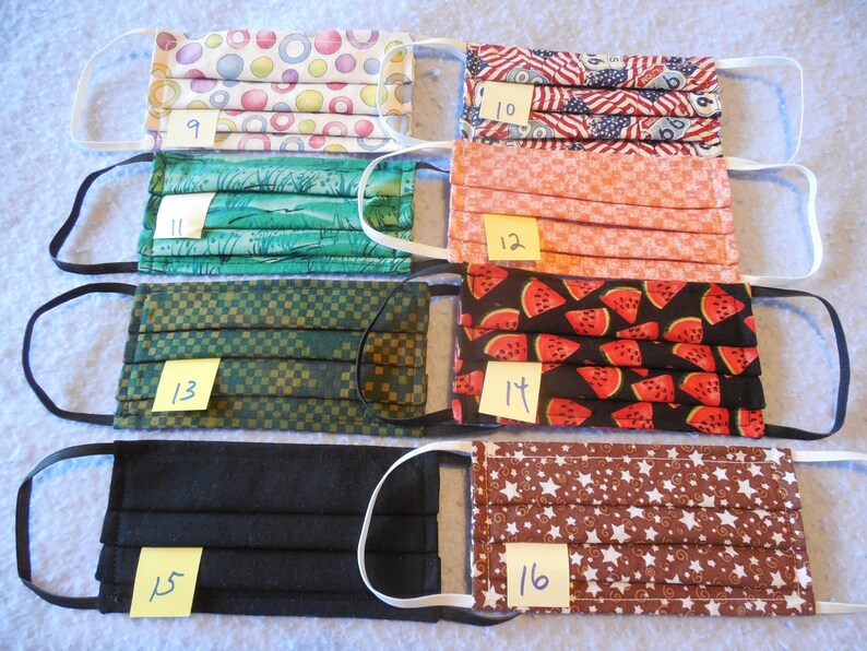 May include: Assortment of pleated face masks with elastic ear loops. Patterns include colorful circles, American flags, a green landscape, a peach-colored grid, watermelon slices, a black solid, and brown stars. Each mask has a number on a yellow sticky note.