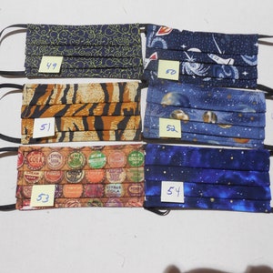 May include: Assortment of six pleated fabric face masks with black elastic ear loops. The masks feature various patterns, including a navy blue swirl design, a tiger stripe print, and a soda bottle cap design. Other masks have space and galaxy themes.