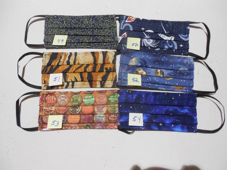May include: Assortment of six pleated fabric face masks with black elastic ear loops. The masks feature various patterns, including a navy blue swirl design, a tiger stripe print, and a soda bottle cap design. Other masks have space and galaxy themes.