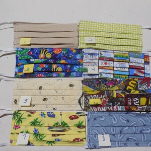 May include: Eight different fabric face masks with various patterns. The masks are all folded and have elastic ear loops. The patterns include beige, green, blue with bees, license plates, bees on a cream background, comic book characters, palm trees and boats on a yellow background, and blue with feathers.