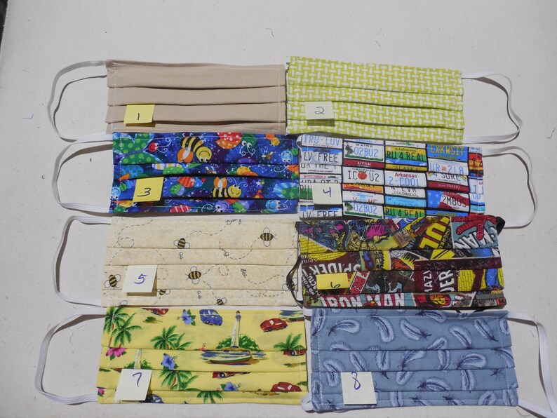 May include: Eight different fabric face masks with various patterns. The masks are all folded and have elastic ear loops. The patterns include beige, green, blue with bees, license plates, bees on a cream background, comic book characters, palm trees and boats on a yellow background, and blue with feathers.