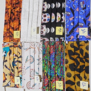 May include: Eight different fabric face masks with various patterns. The masks are all folded and have elastic ear loops. The patterns include a flame design, a white design, a skull design, a US Marine design, a guitar design, a dreamcatcher design, a lizard design, and a star design.