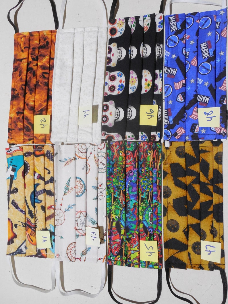 May include: Eight different fabric face masks with various patterns. The masks are all folded and have elastic ear loops. The patterns include a flame design, a white design, a skull design, a US Marine design, a guitar design, a dreamcatcher design, a lizard design, and a star design.