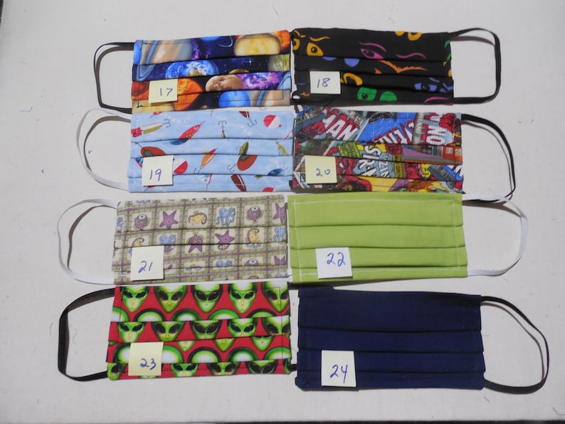 May include: A collection of seven face masks with various patterns and colors. Designs include planets, cartoon eyes, fishing lures, comic book panels, sea creatures, solid green, alien heads, and solid navy blue. Each mask has elastic ear loops.