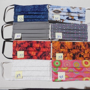 May include: A collection of eight pleated fabric face masks with various patterns. Designs include blue leaves, a navy military theme, solid gray, gray floral, fiery red, orange flames, white textured, and pink floral. Each mask has elastic ear loops and a numbered tag.