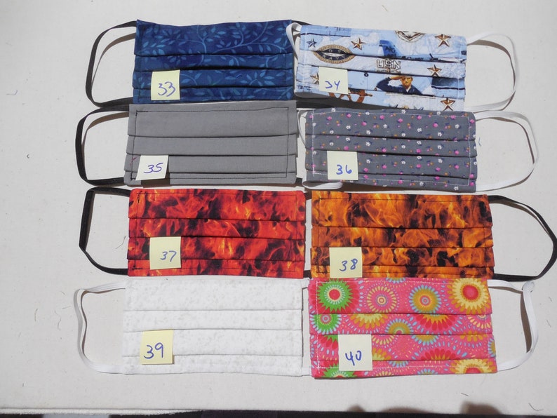 May include: A collection of eight pleated fabric face masks with various patterns. Designs include blue leaves, a navy military theme, solid gray, gray floral, fiery red, orange flames, white textured, and pink floral. Each mask has elastic ear loops and a numbered tag.