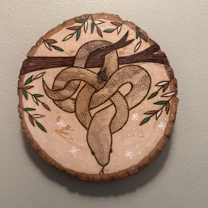 May include: A circular wooden art piece featuring a detailed illustration of a snake intertwined around a branch. The snake is tan with a textured pattern. The artwork includes green and brown leaves and white snowflake accents.