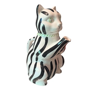 May include: A ceramic teapot shaped like a seated cat with a white base and black vertical stripes. The cat features light green eyes and a light green collar around its neck. The spout extends from the cat's chest, and a handle is on the back.