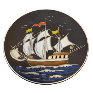 May include: A round decorative plate featuring a dark background with a sailing ship illustration. The ship has white sails, a brown hull, and yellow and orange flags. Below the ship, stylized blue and white waves represent the ocean.