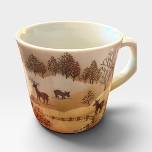 May include: A ceramic mug featuring a wrap-around illustration of a woodland scene with brown deer, bare trees, and a fence. The background consists of soft tan and white tones, depicting a landscape with autumn-colored foliage and a clear handle.