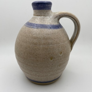 May include: A handmade stoneware jug with a rounded body and a handle. The jug is primarily beige with a blue band around the middle and the top. The jug is likely used for holding liquids and has a rustic, artisanal appearance.