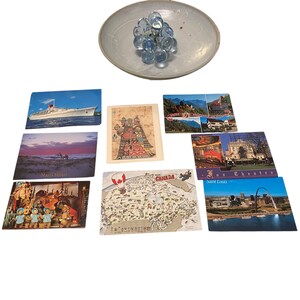 May include: A collection of vintage postcards featuring various travel destinations and a decorative glass orb cluster in a shallow, light blue dish. Postcards depict landscapes, architecture, and a cruise ship.