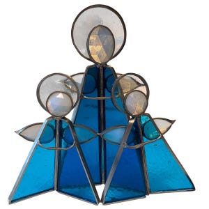 Puede incluir: Una escultura de ángel de vidrieras con tres figuras en tonos azules y vidrio transparente. Cada figura tiene un halo y alas, siendo la figura central la más grande. El vidrio está engastado en un marco de metal.