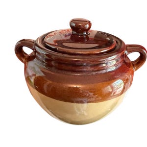 May include: A small, round, ceramic pot with a lid. The pot is primarily brown with a lighter tan band around the base. It has two handles and a knob on the lid. The pot appears to be glazed and has a glossy finish.