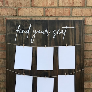 May include: A dark brown wooden sign with the text "find your seat" written in white cursive. The sign has a string of twine running across it with white cards attached with clothespins.