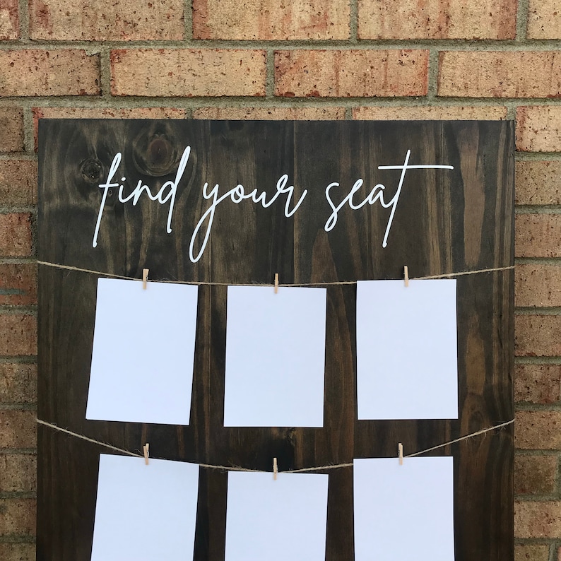 May include: A dark brown wooden sign with the text "find your seat" written in white cursive. The sign has a string of twine running across it with white cards attached with clothespins.