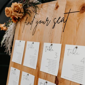 May include: A wooden sign with the text "find your seat" written in black cursive. The sign is decorated with white cards with black cursive text indicating table numbers and guest names. The sign is decorated with dried flowers.