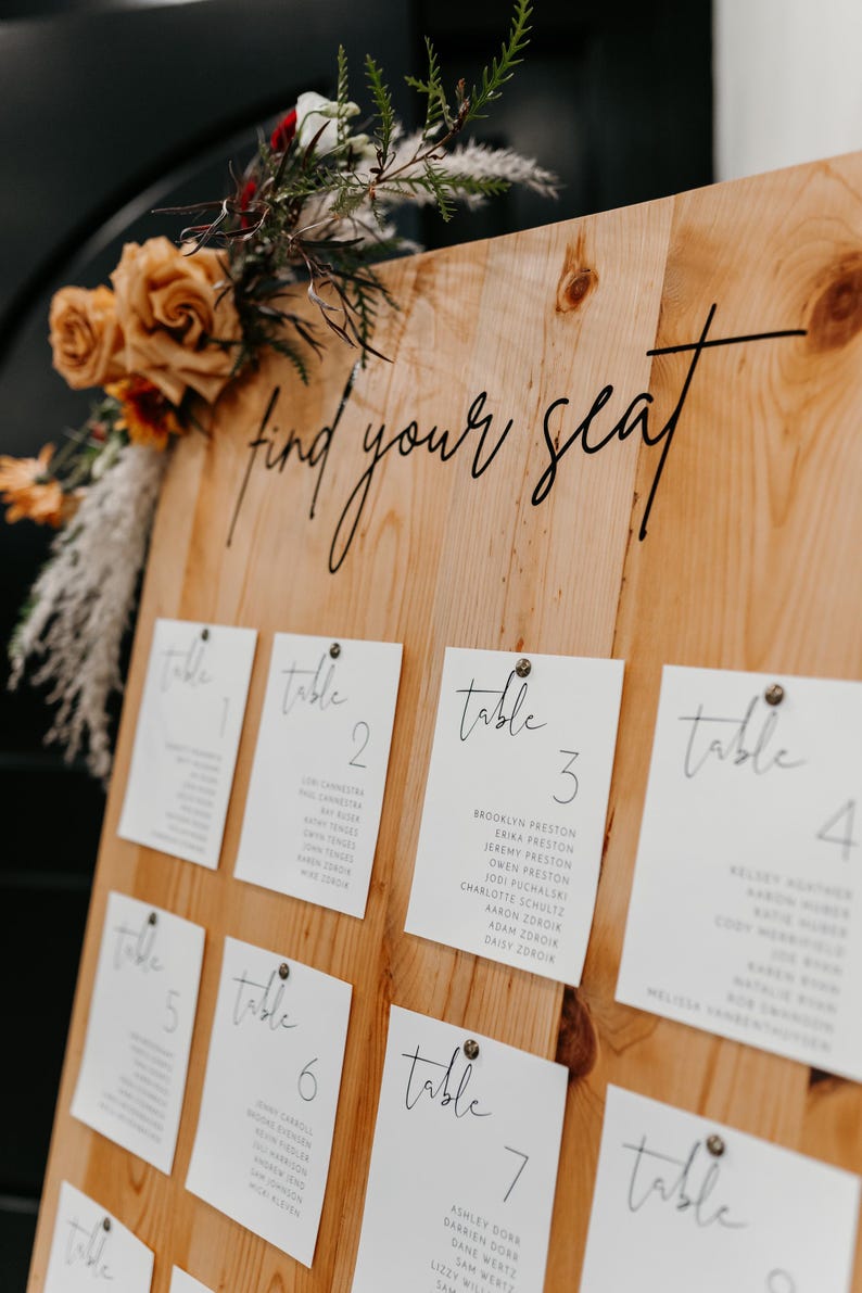 May include: A wooden sign with the text "find your seat" written in black cursive. The sign is decorated with white cards with black cursive text indicating table numbers and guest names. The sign is decorated with dried flowers.