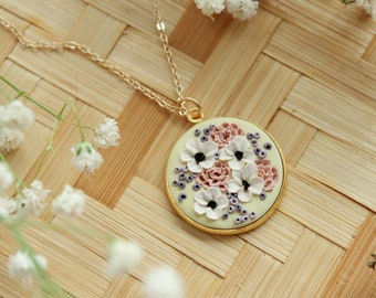 May include: A gold chain necklace with a round pendant featuring a floral design. The pendant is white with pink and purple flowers and a light green background.