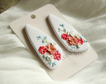 May include: A pair of white ceramic earrings with a floral design. The flowers are pink, red, and white with blue accents. The earrings are shaped like a teardrop.