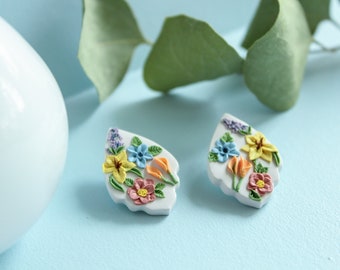 Op de afbeelding: Een paar witte keramische oorbellen met een bloemenmotief. De bloemen zijn in tinten geel, oranje, roze, blauw en paars.