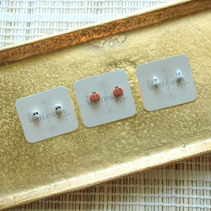May include: Three pairs of Halloween-themed stud earrings on white cards, displayed on a gold tray. The earrings feature a skull, a pumpkin, and a ghost design. The cards have a watermark.