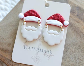 May include: Santa Claus-themed earrings. Each earring features a red glittery Santa hat with a white trim and a small white pom-pom. Below the hat is a white beard with a mustache. The earrings are attached to a white card with the text "the WATERMARK design".