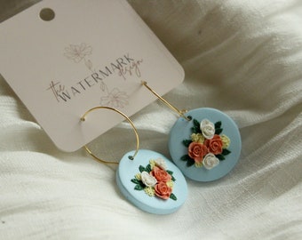 May include: A pair of gold hoop earrings with light blue round charms featuring a floral design with white and peach flowers. The earrings are on a white fabric background with a tag that says "The Watermark Design".