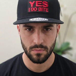 May include: Black flat-brimmed baseball cap with the text "YES I DO BITE" in red embroidery. The cap has a square patch with the text and a white tag. The cap is worn by a person.