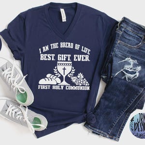 May include: Navy blue V-neck t-shirt with the text "I AM THE BREAD OF LIFE BEST. GIFT. EVER." and "FIRST HOLY COMMUNION" in white. The shirt is paired with blue jeans and gray sneakers.