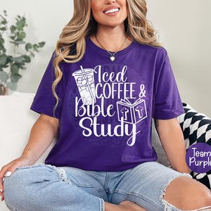 May include: A purple t-shirt with the text "Iced Coffee & Bible Study" in white script. The design includes a graphic of an iced coffee and an open book with a cross. The shirt is a solid purple color.