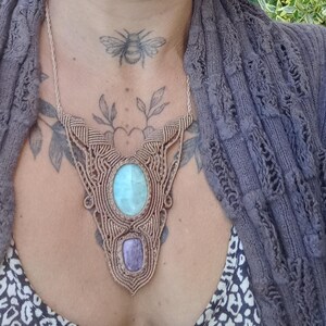 May include: A tan macrame necklace featuring a large, light blue oval gemstone and a smaller, purple rectangular gemstone. The necklace is worn over a patterned top and a grey knitted cardigan. A bee tattoo is visible on the neck.