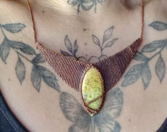 May include: A handmade macrame necklace featuring a large, oval-shaped, light green gemstone pendant. The necklace has a brown macrame design and a delicate chain. The image shows the necklace worn on a person's chest.