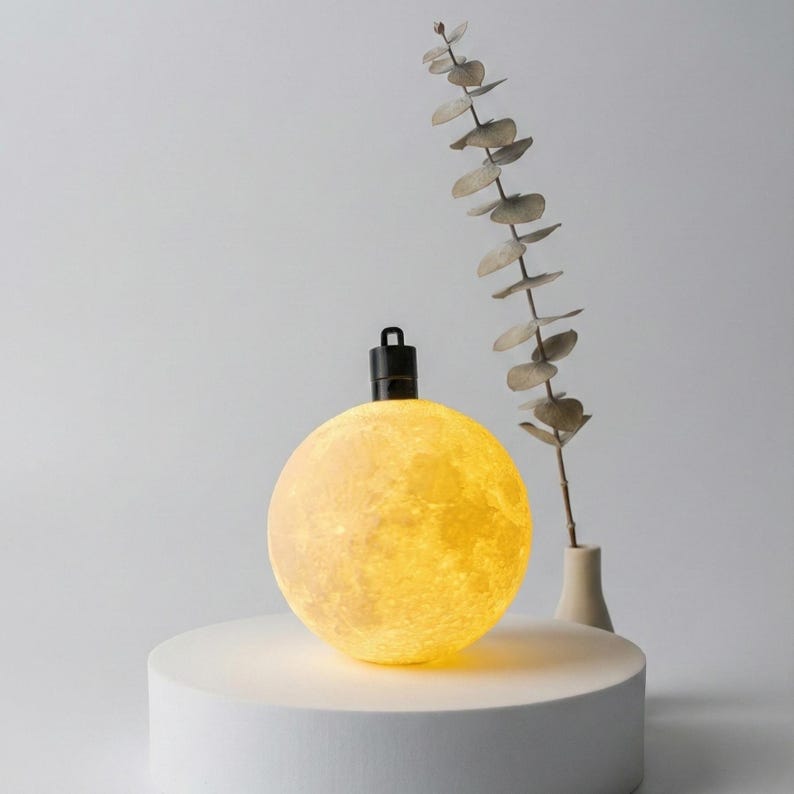 Minimalist display of a glowing mini moon ornament on a clean white pedestal. A single dried botanical stem stands nearby against a soft grey background. Highlights the warm light and detailed crater texture.