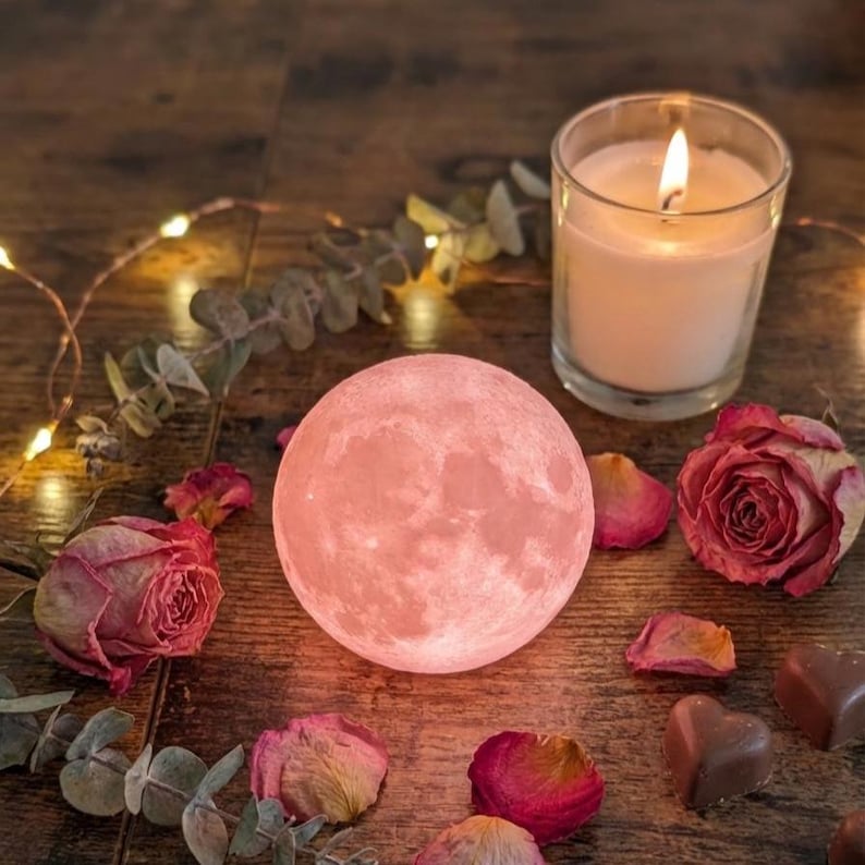 Glowing pink 3D printed moon ornament on a cozy wooden surface with soft Valentine’s lighting, surrounded by romantic decor like dried flowers or a candle. Product centered and glowing with realistic lunar detail.