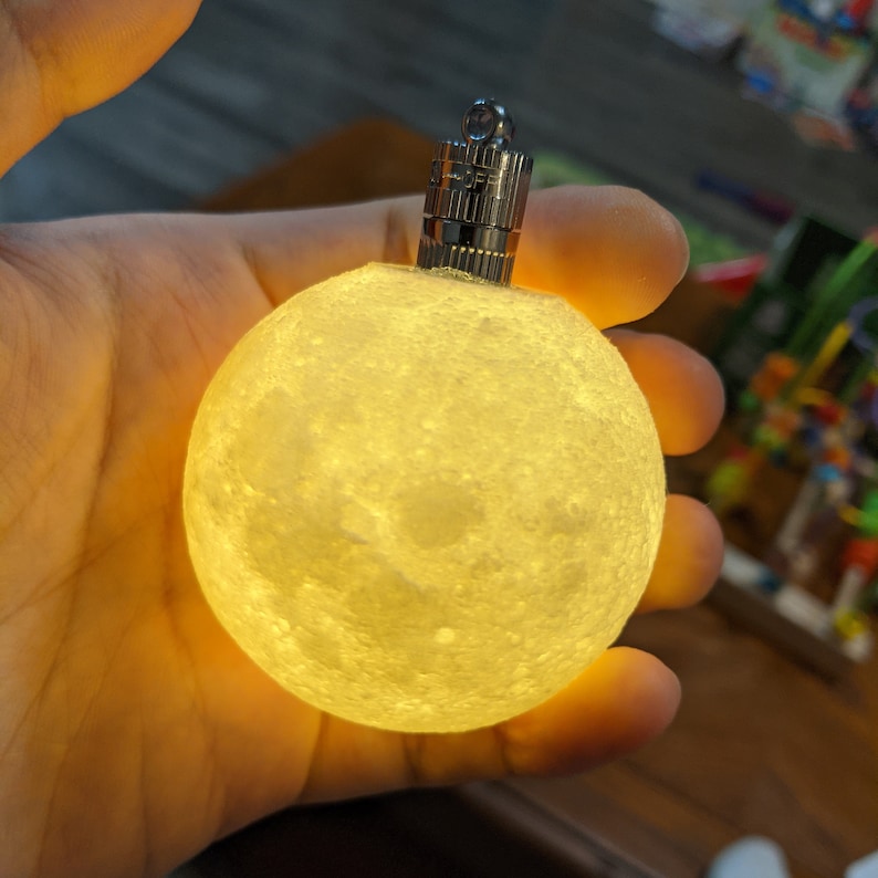 May include: A glowing, spherical moon lamp with a textured surface, held in a hand. The lamp emits a soft, warm light and has a silver metal top with an on/off switch. The lamp is a decorative item.