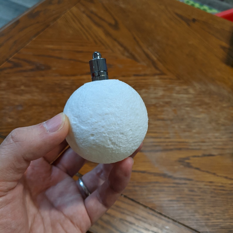 May include: A white, textured sphere Christmas ornament with a silver metal cap. The ornament is held in a hand against a wooden background. The sphere has a rough, porous surface.