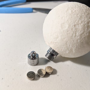 May include: A white, textured sphere with a silver metal base, resembling the moon. Beside it are a silver metal cap and three small, round batteries. The items are on a white surface.